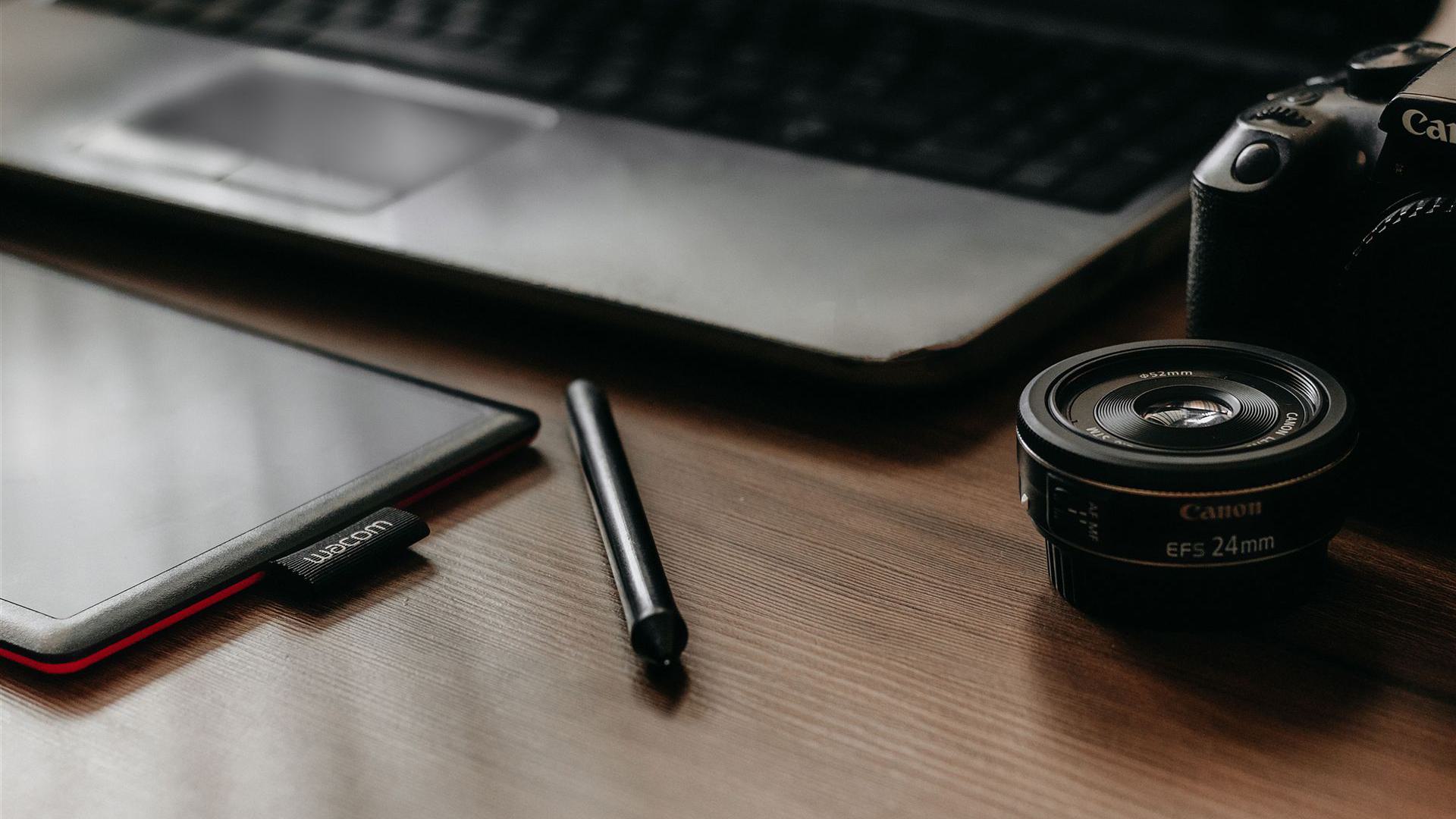 Ein Arbeitsplatz mit Laptop, Tablet, Kamera und Objektiv, ideal für kreative Arbeiten und Fotografie.